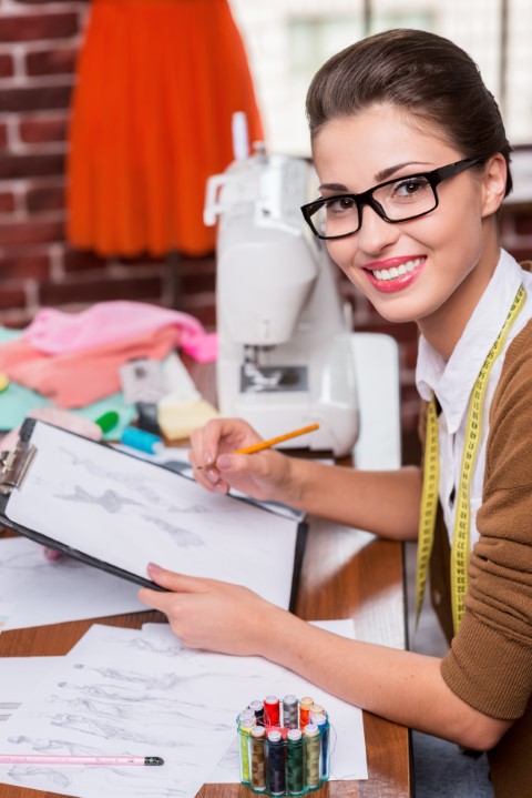 Drawing fashion sketch. Top view of beautiful young fashion designer drawing a fashion sketch and smiling at camera while sitting at her working place